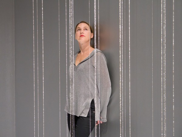 Eine Frau steht in einem Raum mit grauen Wänden, durch den vertikale, schimmernde Fäden oder Lichtstränge verlaufen. Sie trägt ein silbergraues Oberteil und blickt leicht nach oben. Die Atmosphäre wirkt ruhig und konzentriert, das Licht reflektiert an den glänzenden Strängen.