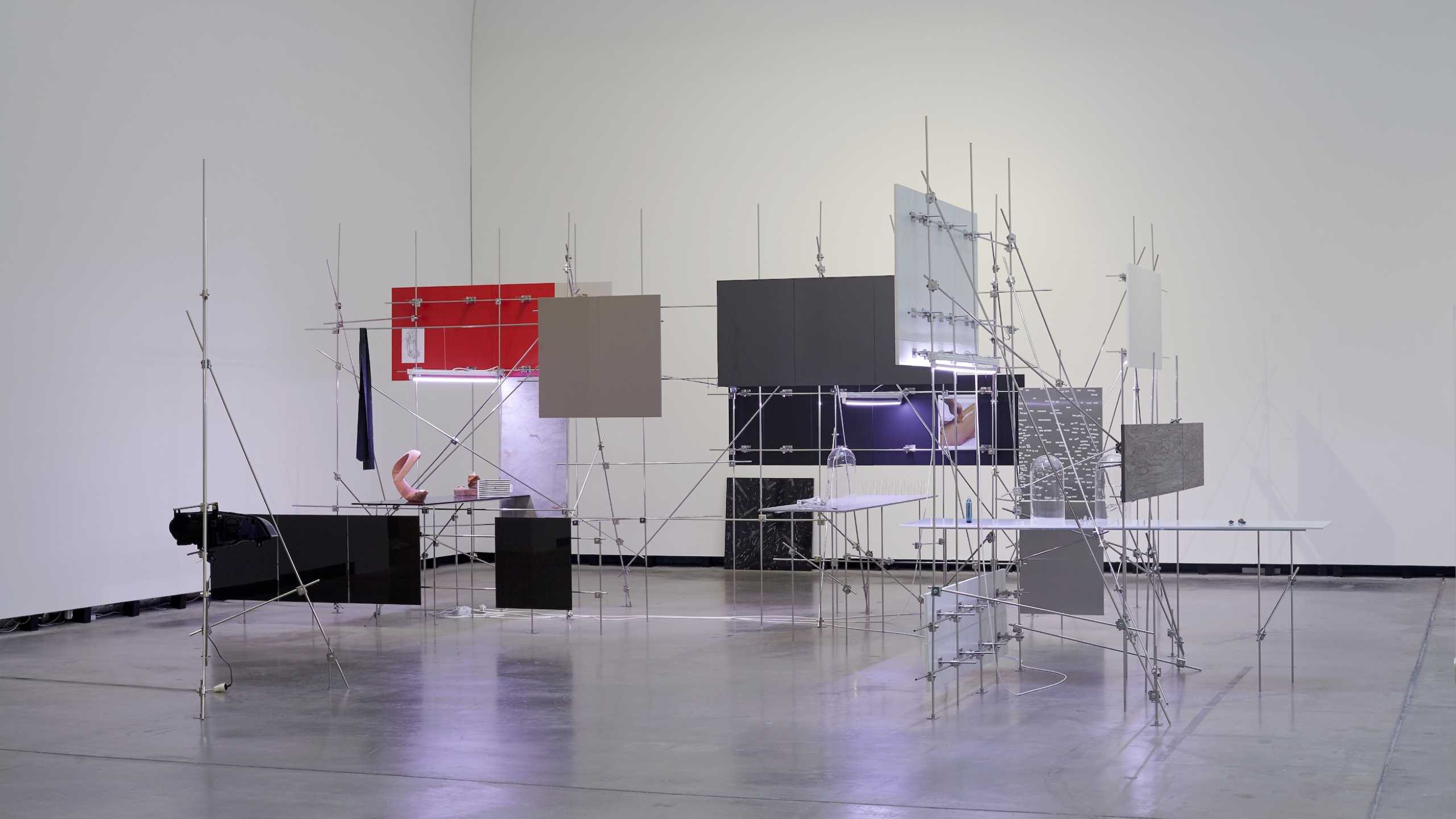 Komplexe Kunstinstallation in einem weißen Ausstellungsraum. Die Struktur besteht aus filigranen Metallgerüsten, die mit rechteckigen Tafeln in verschiedenen Farben und Materialien bestückt sind, darunter Rot, Schwarz, Grau und transluzente Flächen. Dazwischen sind kleine Objekte arrangiert, wie Glasglocken und modellhafte Körperteile. Die Installation wirkt technisch, experimentell und hat einen skulpturalen Charakter.“
