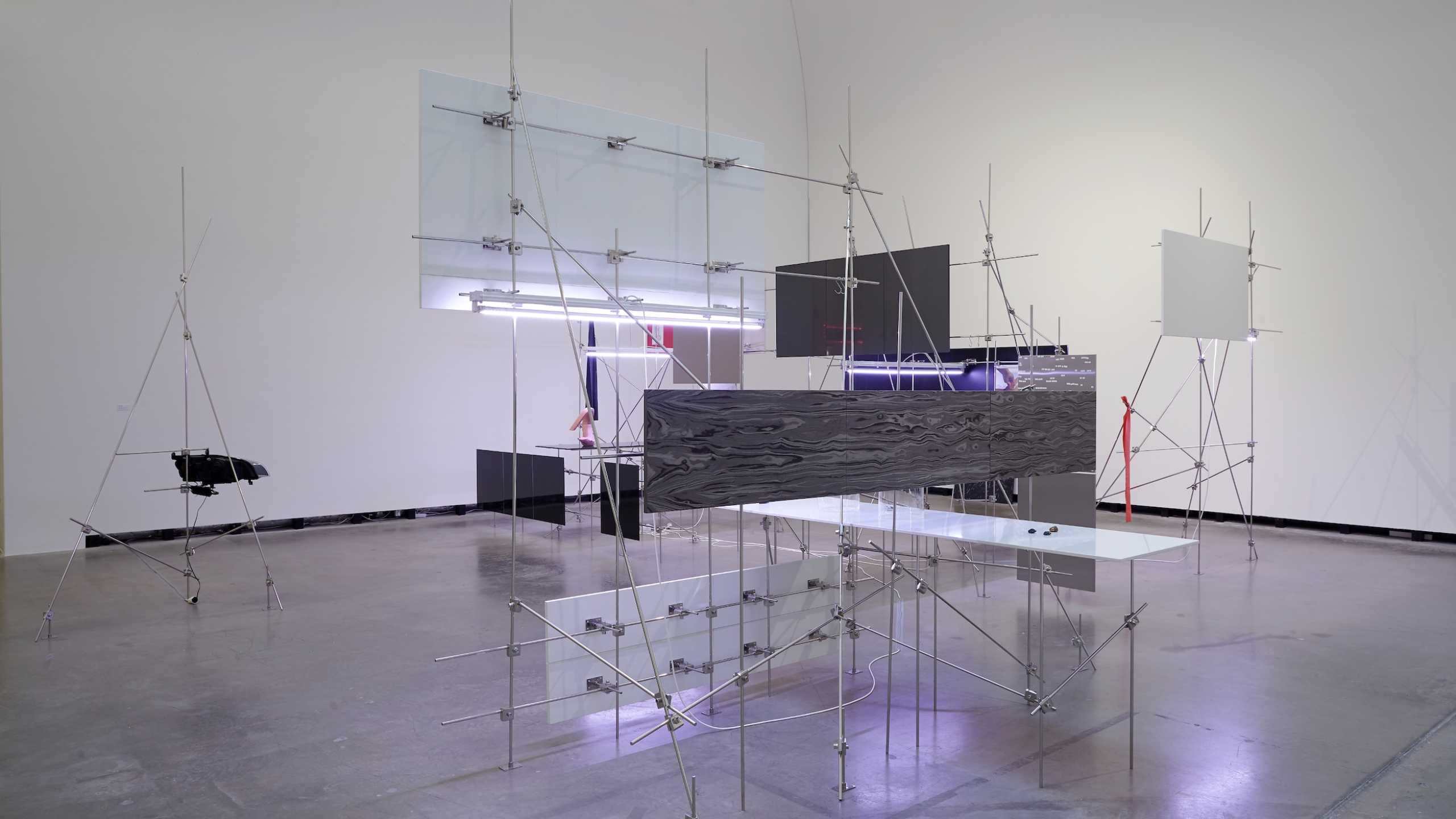 Kunstinstallation in einem weißen Ausstellungsraum. Die Installation besteht aus filigranen Metallgerüsten, die durch Stangen und Knoten verbunden sind. Daran sind verschiedenfarbige rechteckige Flächen aus Glas, Kunststoff oder Metall befestigt. Einige Flächen sind transluzent, andere schwarz oder in Marmoroptik. Die Struktur wirkt technisch und abstrakt, mit einem industriellen Charakter.