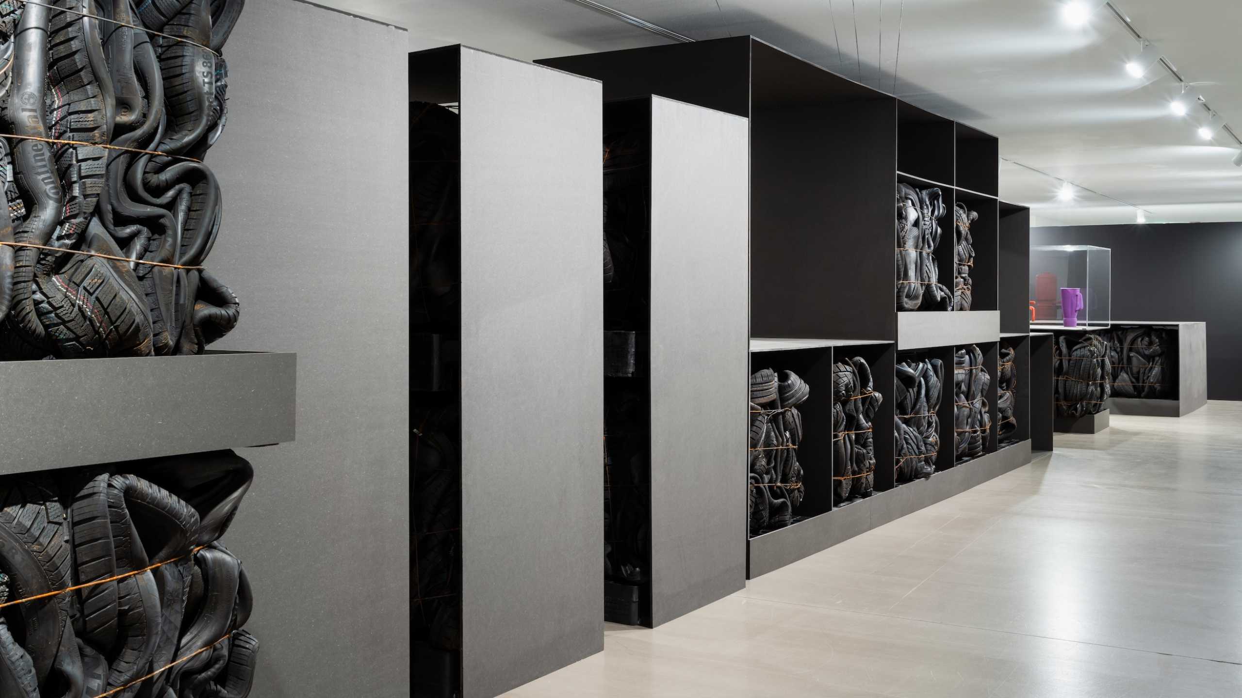 Kunstinstallation in einem modern beleuchteten Raum. In offenen Regalen und Nischen sind gebrauchte, verformte Autoreifen zu Blöcken zusammengeschnürt und gestapelt. Die Reifen sind schwarz und wirken durch die Enge der Schnürung wie organische Skulpturen. Rechts im Hintergrund steht eine Vitrine mit farbigen Objekten. Die Raumgestaltung ist minimalistisch mit grauen und schwarzen Flächen sowie poliertem Betonboden.