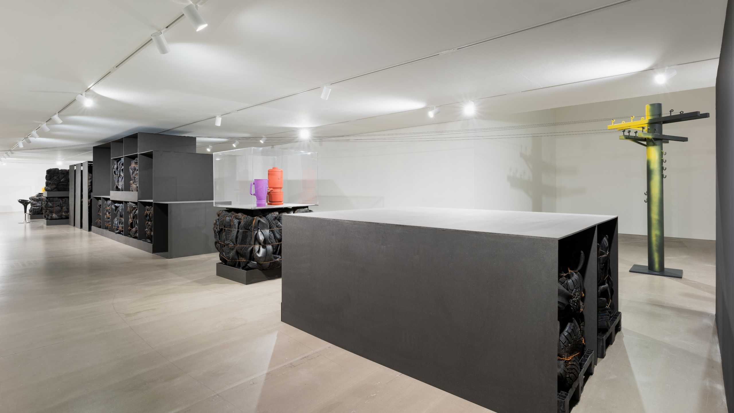 Ausstellungsraum mit einer großformatigen Kunstinstallation. Im Raum stehen mehrere schwarze Regalkonstruktionen, in denen gebrauchte Autoreifen gepresst und gestapelt sind. In der Mitte befindet sich ein rechteckiger schwarzer Block, ebenfalls mit Reifenmodulen gefüllt. Rechts vorne steht eine Vitrine auf einem Reifenblock, darin befinden sich bunte Objekte, darunter ein oranger und ein violetter Zylinder. Im Hintergrund steht eine gelb-schwarz gefärbte Skulptur in Form eines stilisierten Strommastes