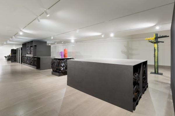 Ausstellungsraum mit einer großformatigen Kunstinstallation. Im Raum stehen mehrere schwarze Regalkonstruktionen, in denen gebrauchte Autoreifen gepresst und gestapelt sind. In der Mitte befindet sich ein rechteckiger schwarzer Block, ebenfalls mit Reifenmodulen gefüllt. Rechts vorne steht eine Vitrine auf einem Reifenblock, darin befinden sich bunte Objekte, darunter ein oranger und ein violetter Zylinder. Im Hintergrund steht eine gelb-schwarz gefärbte Skulptur in Form eines stilisierten Strommastes