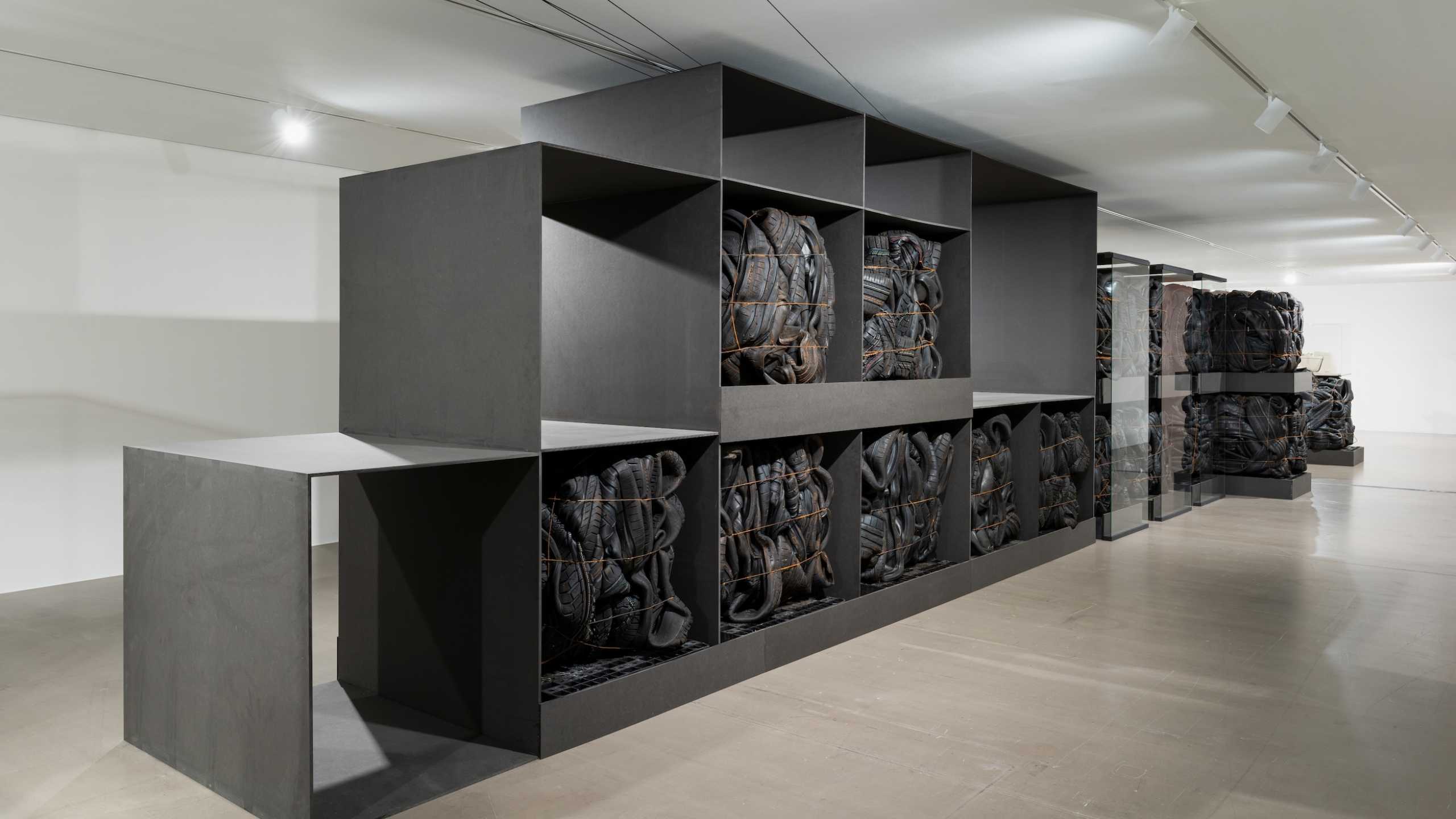 Kunstinstallation in einem hellen Ausstellungsraum mit minimalistischer Ästhetik. In großen, schwarzen Regalelementen sind eng zusammengeschnürte, gebrauchte Autoreifen gestapelt. Die Reifen sind in jedem Fachblock dicht gepackt und mit Seilen fixiert, wodurch sie wie kompakte, plastische Module wirken. Der Boden besteht aus glattem, hellem Beton, und die Decke ist mit einer schlichten Lichtschiene ausgestattet.