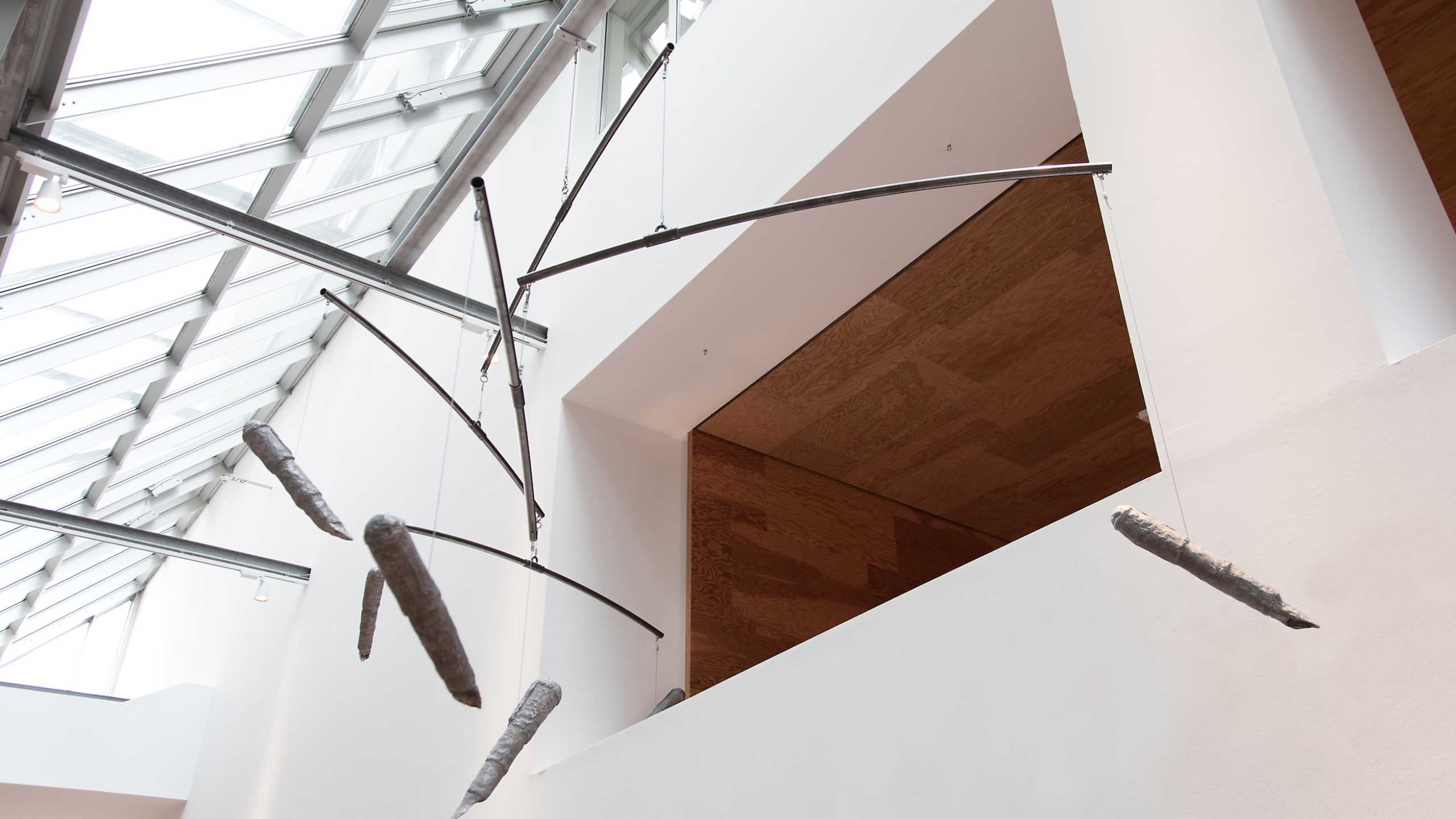 Moderne Hängeskulptur in einem lichtdurchfluteten Raum mit Glasdach. Die Skulptur besteht aus gebogenen Metallstangen, die an dünnen Drähten von der Decke hängen. Daran befestigt sind längliche, unregelmäßig geformte Objekte, die wie in Stoff eingewickelt wirken. Die Gesamtform erinnert an ein Mobile, das sich sanft im Raum bewegen könnte.