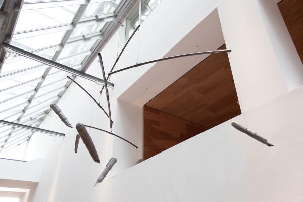 Moderne Hängeskulptur in einem lichtdurchfluteten Raum mit Glasdach. Die Skulptur besteht aus gebogenen Metallstangen, die an dünnen Drähten von der Decke hängen. Daran befestigt sind längliche, unregelmäßig geformte Objekte, die wie in Stoff eingewickelt wirken. Die Gesamtform erinnert an ein Mobile, das sich sanft im Raum bewegen könnte.