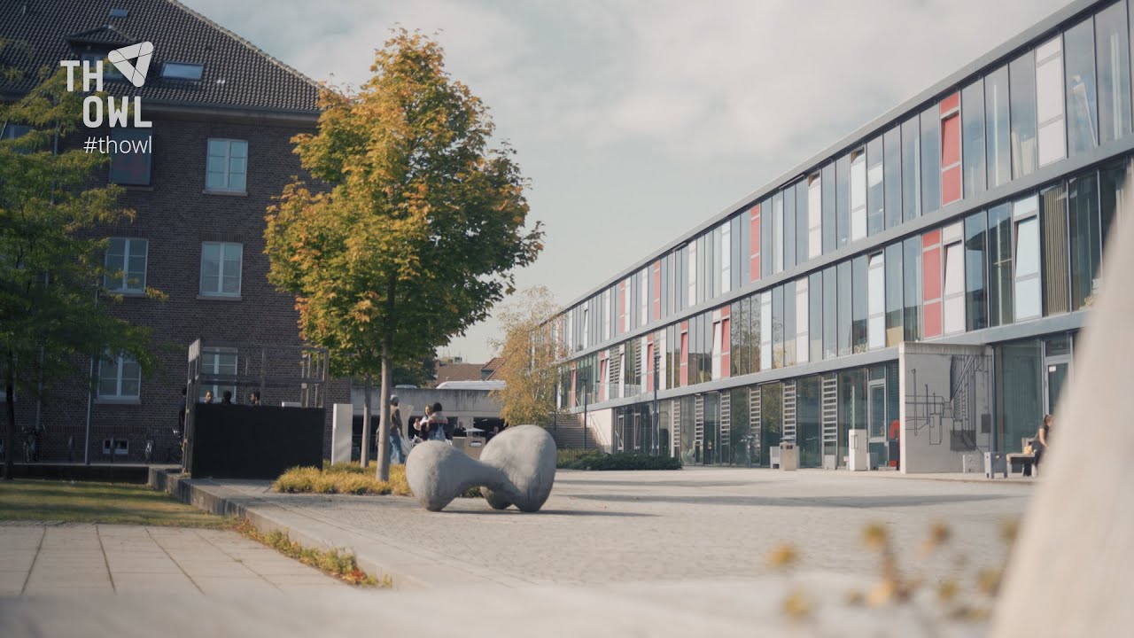 Innenhof einer Hochschule mit modernen und historischen Gebäuden. Rechts ein langgestrecktes, modernes Gebäude mit großen Glasflächen und farbigen Fensterelementen. Links ein älteres Backsteingebäude mit Satteldach. Dazwischen Bäume mit herbstlich gefärbten Blättern, ein geschwungenes Betonkunstwerk auf dem Platz, und Studierende, die sich im Freien aufhalten. Im oberen linken Bildbereich ist das Logo der Technischen Hochschule Ostwestfalen-Lippe (TH OWL) sichtbar.