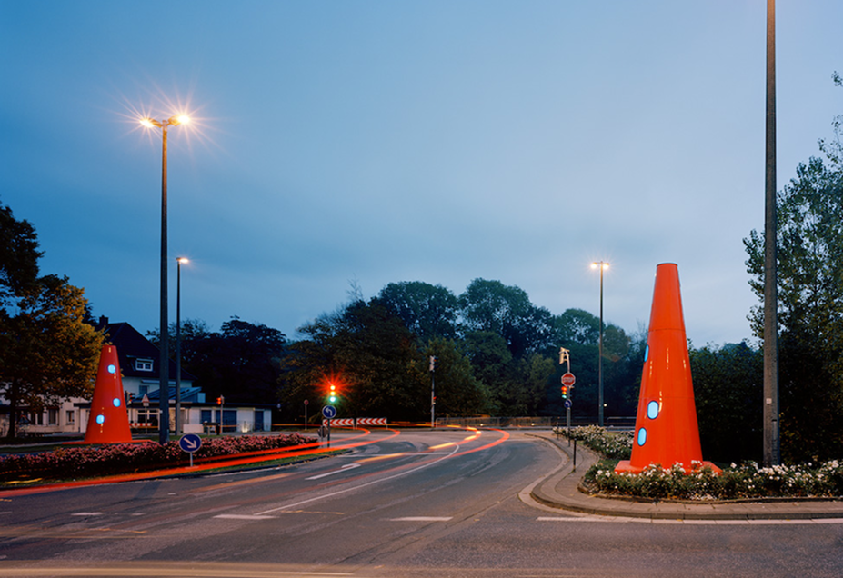 Eine nächtliche Straßenszene mit zwei großen, orangefarbenen, skulpturalen Kegeln mit blauen, runden Fenstern, die an Verkehrsleitkegel erinnern. Sie stehen in einem bepflanzten Kreisverkehr, umgeben von Straßenlaternen und vorbeiziehenden Lichtspuren von Autos.